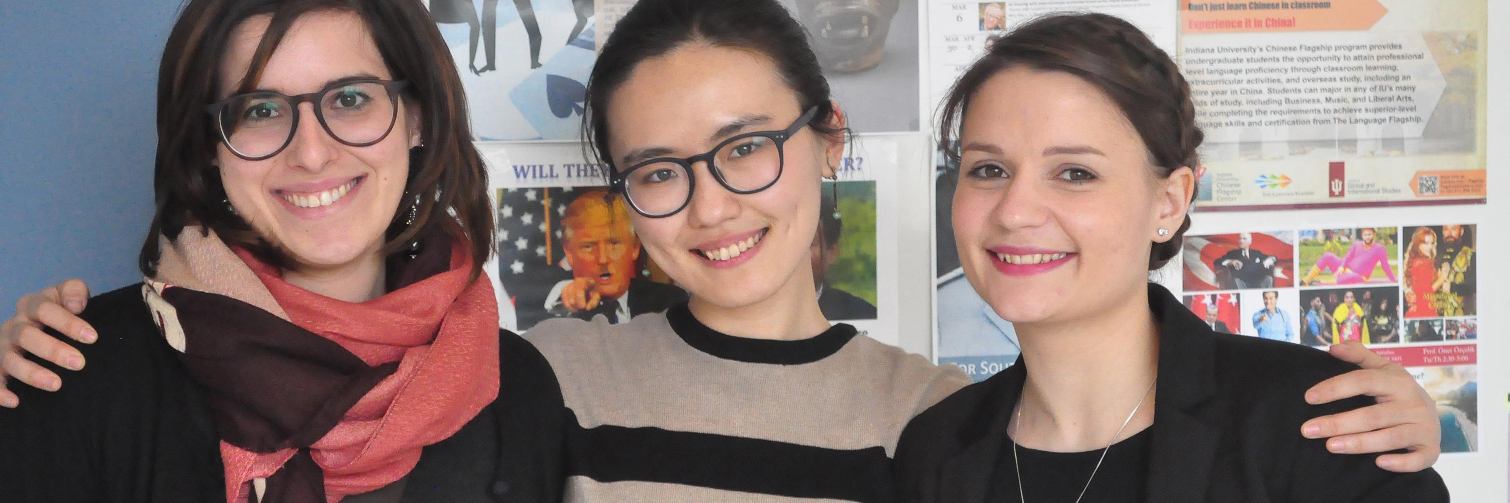 Three Indiana University French and Italian graduate students pose for a photo in front of a classroom bulletin board.
