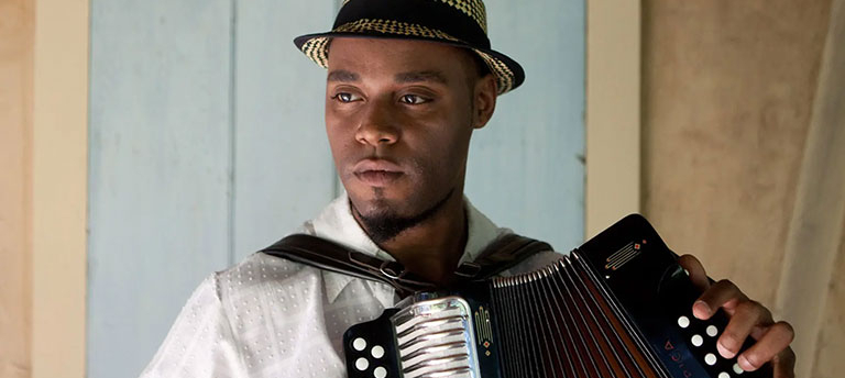 A headshot of Cedric Watson, who wears a white shirt and holds an accordion.