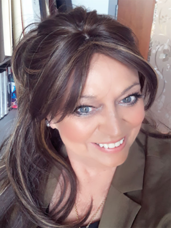 A headshot of Connie May, who wears a brown blazer and poses near a bookshelf.