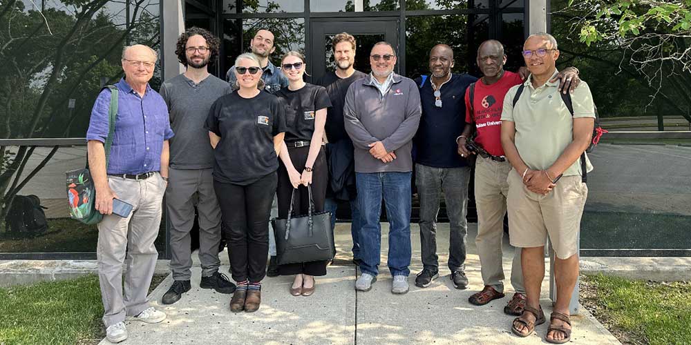 A group of ten people stands smiling outside a modern glass building on a sunny day.
