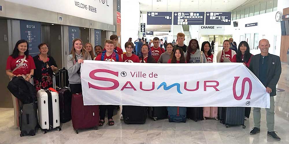 A large group of students and chaperones hold a “Ville de Saumur” banner in an airport arrivals area, surrounded by luggage.